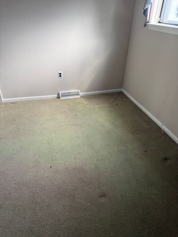 Empty room with stained carpet and beige walls. A small vent is visible.