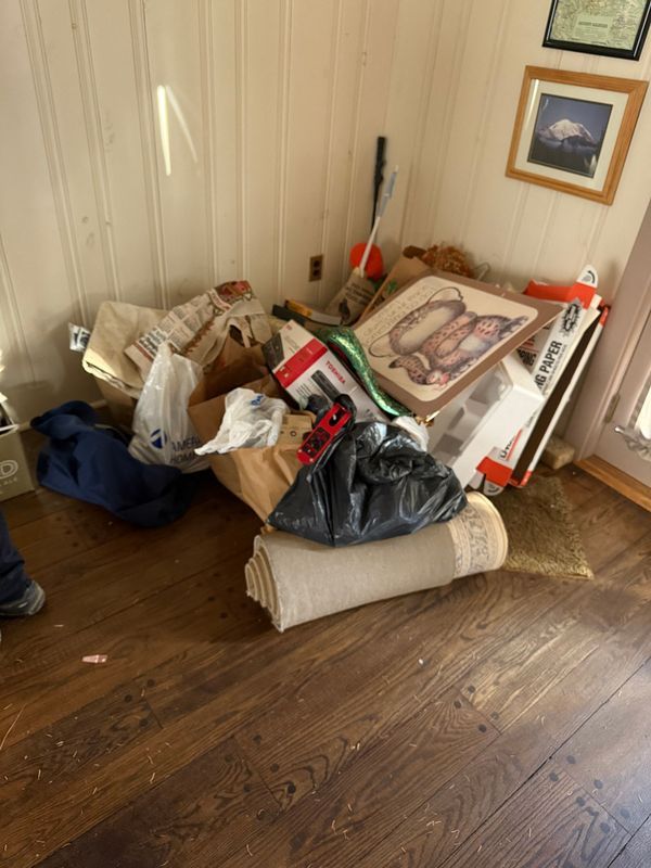 Pile of miscellaneous items against a white wall and wood floor. Includes a rolled-up rug, bags, and boxes.