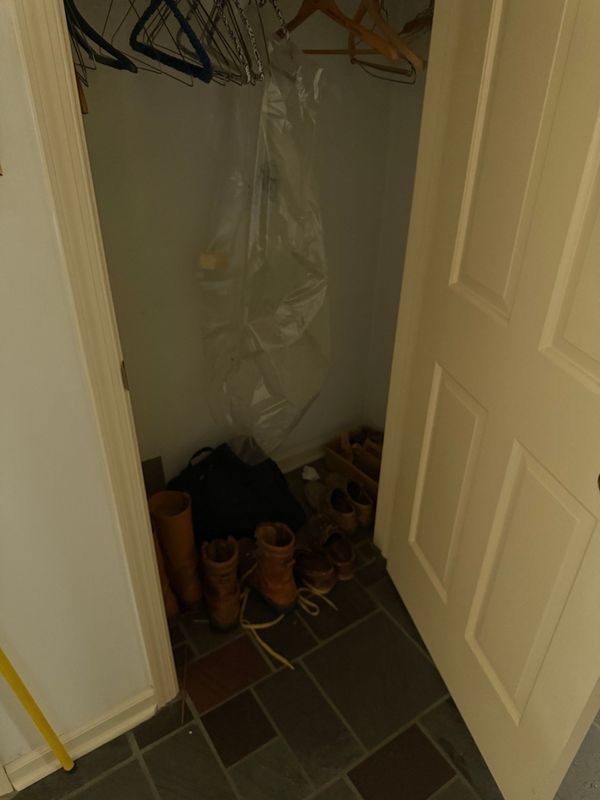 A small, open closet with a few pairs of boots and a garment bag on a dark tile floor.