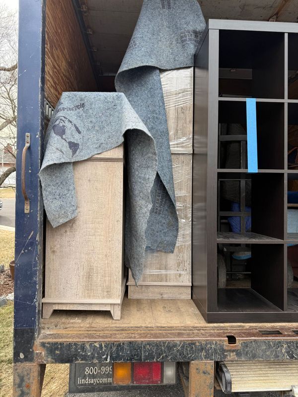 Inside a truck: packed furniture includes a light cabinet, wrapped boxes, and a dark cube shelf, covered in moving blankets.