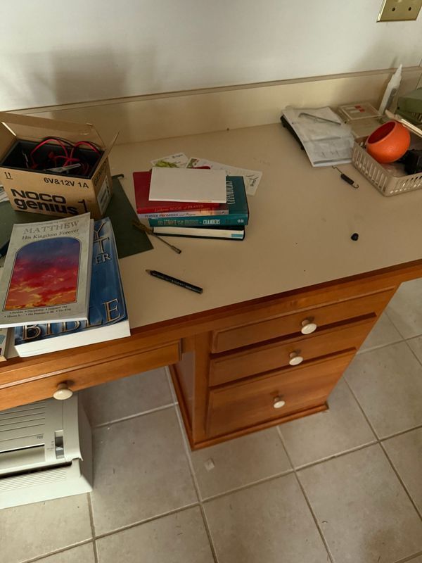 A wooden desk cluttered with books, papers, and a printer. A wall and tiled floor are in the background.