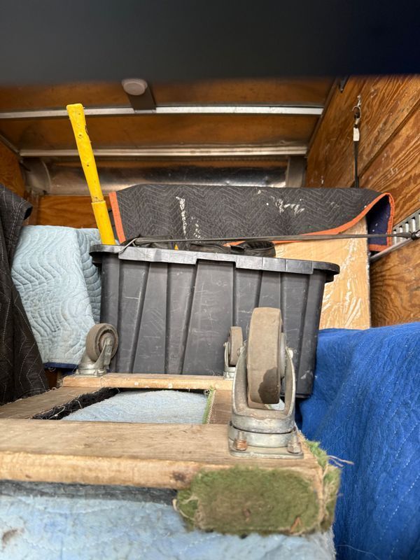 Inside a cargo van: a black bin on a wooden platform with wheels, various tools, and wrapped items.