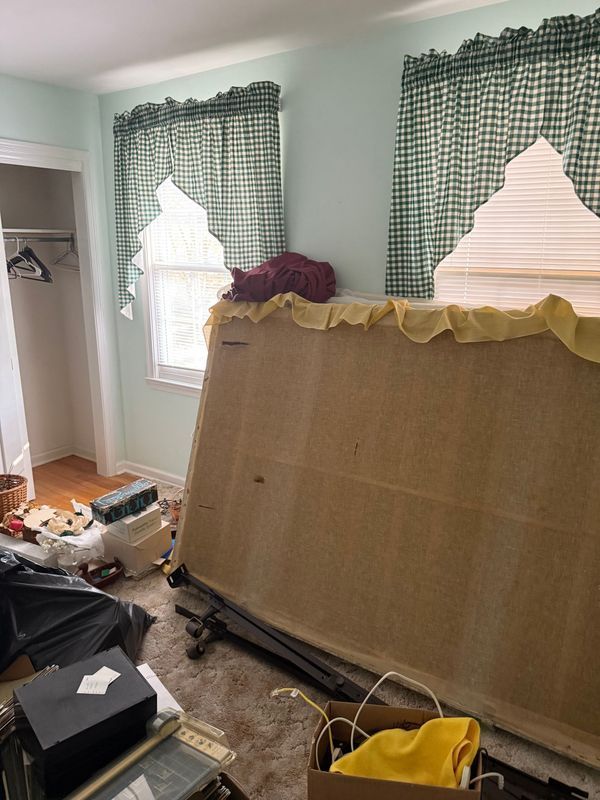 Bedroom with unmade bed, window with green gingham curtains, and cluttered floor.