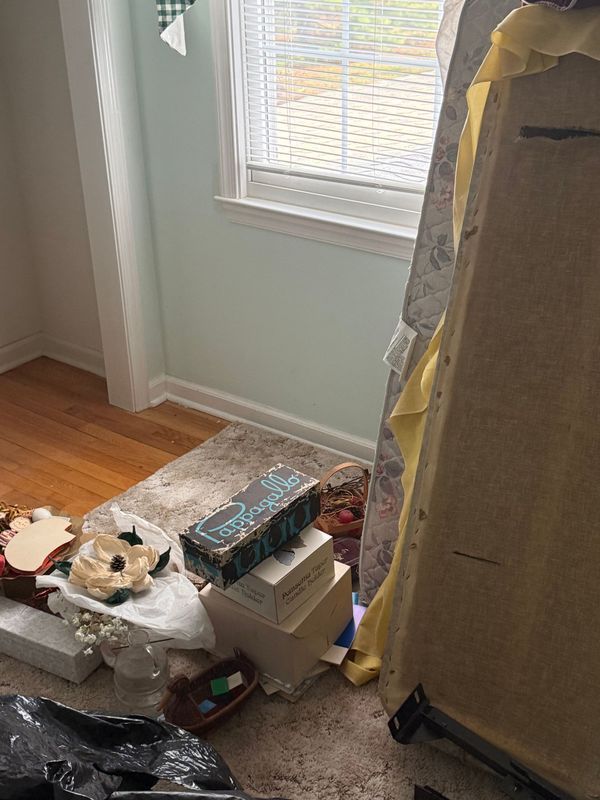 A cluttered corner of a room with a window. Items including a headboard, boxes, and decor sit on the floor.