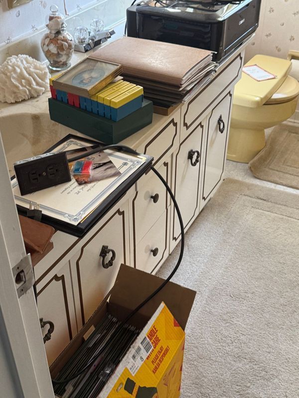 Cluttered bathroom: cabinet with objects on top, toilet, rug, and open cardboard box on floor.