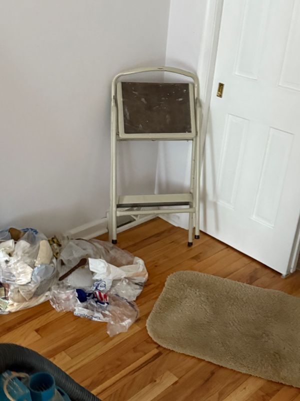A folding step stool in a corner next to a white door. Crumpled trash and a beige bath mat are on the hardwood floor.