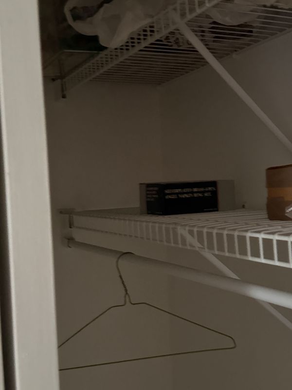 A closet interior with white wire shelving. A hanger hangs below a shelf holding a box and a cylindrical object.