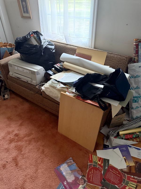 Couch covered in papers, supplies, and a trash bag, in a room with a window and pink carpet.