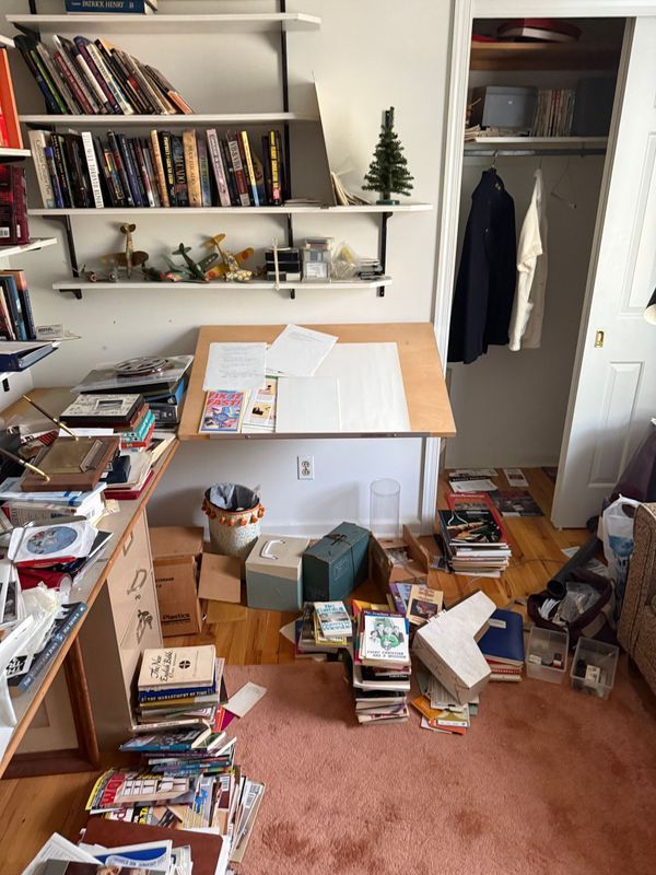 Messy room with drawing table, shelves of books, and cluttered floor, possibly a home office.