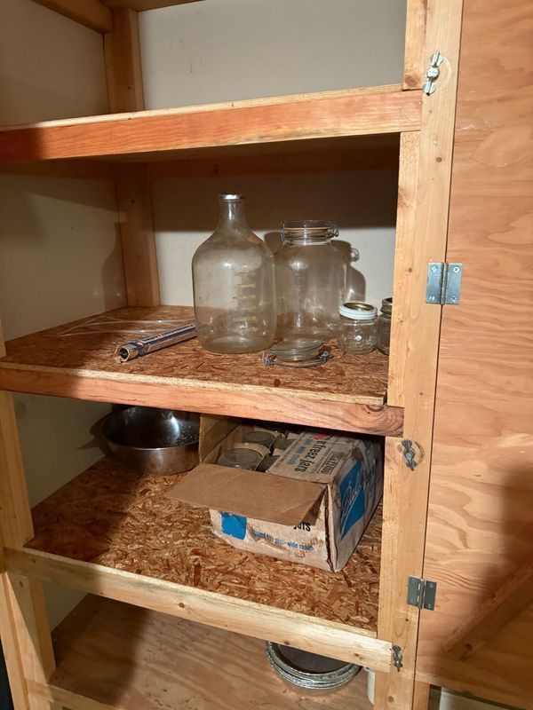 Wooden pantry with shelves. Contains glass jars, metal bowls, and cardboard boxes.