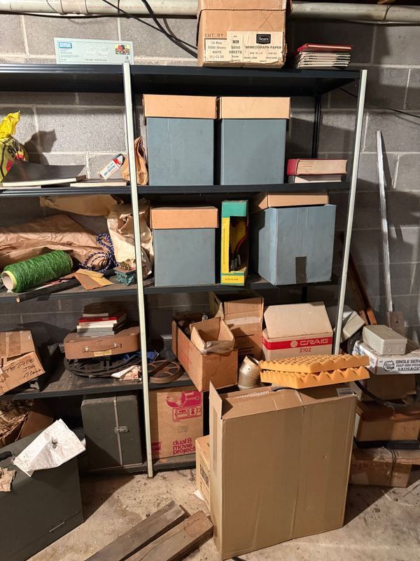 Metal shelves in a basement hold boxes, books, and other objects against a concrete block wall.