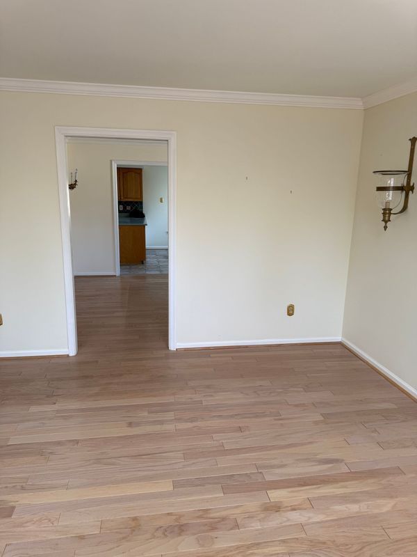 Empty room with light wood floors, doorway to a kitchen, and sconce on the wall.