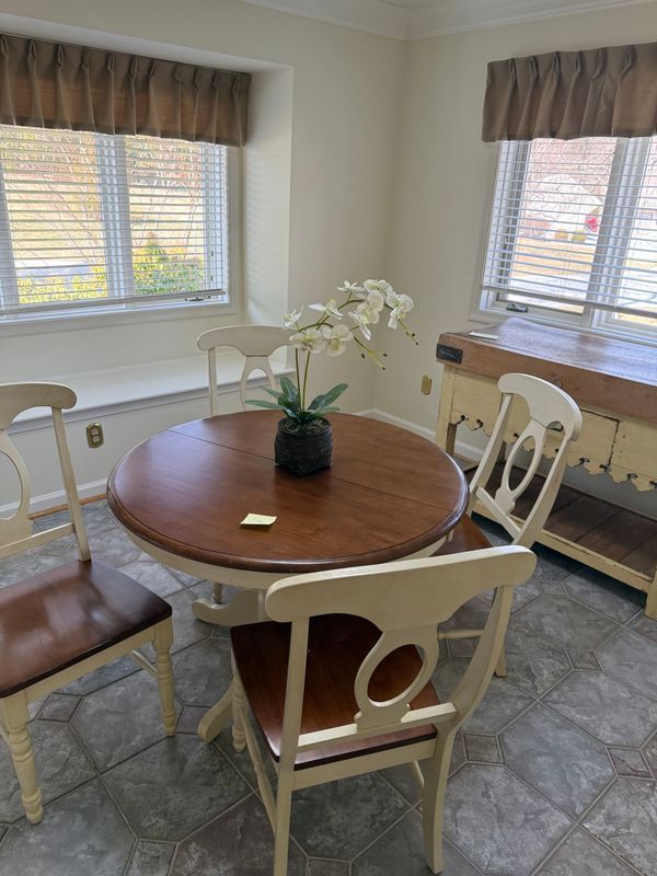 Dining room with a round wooden table, four chairs, and a sideboard. Windows with blinds.