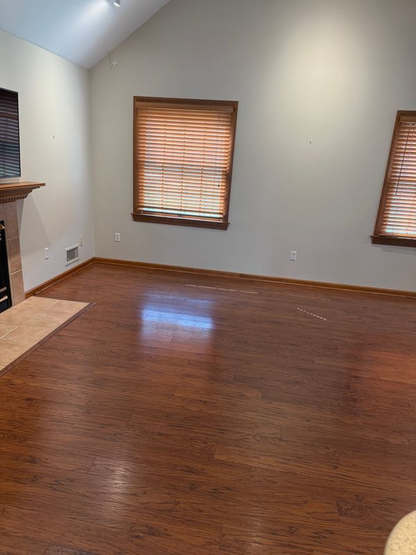 Empty living room with wood floors, two windows with blinds, and a fireplace.