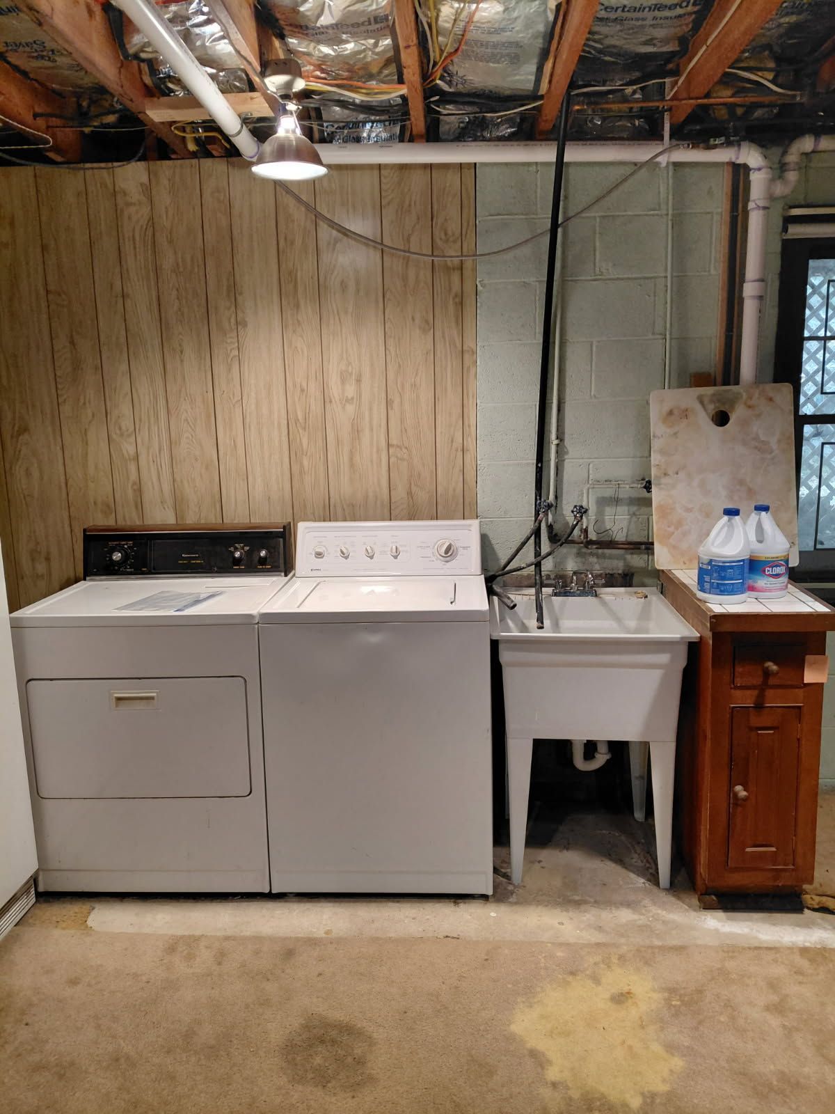 Washer, dryer, and utility sink in a basement laundry area. Two bleach bottles on a wooden cabinet.
