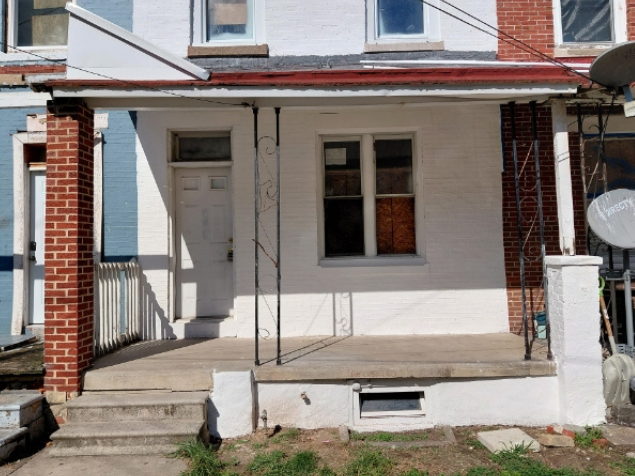 A small, white house with a porch and brick columns. A door and window are visible.