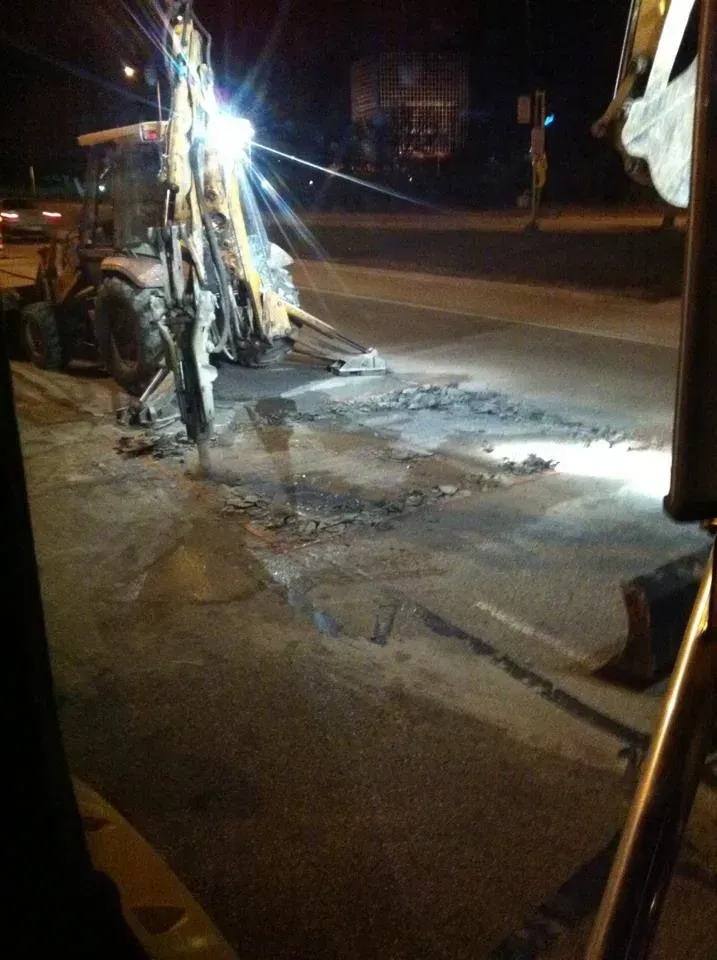 Backhoe removing asphalt on a dark road at night; bright lights illuminate the scene.