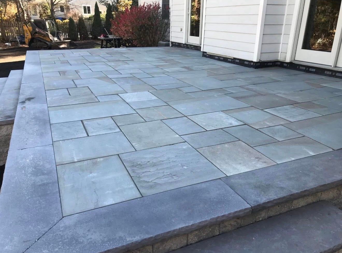 A patio with gray rectangular stone pavers, featuring a matching stone border and stairs, adjacent to a white house.
