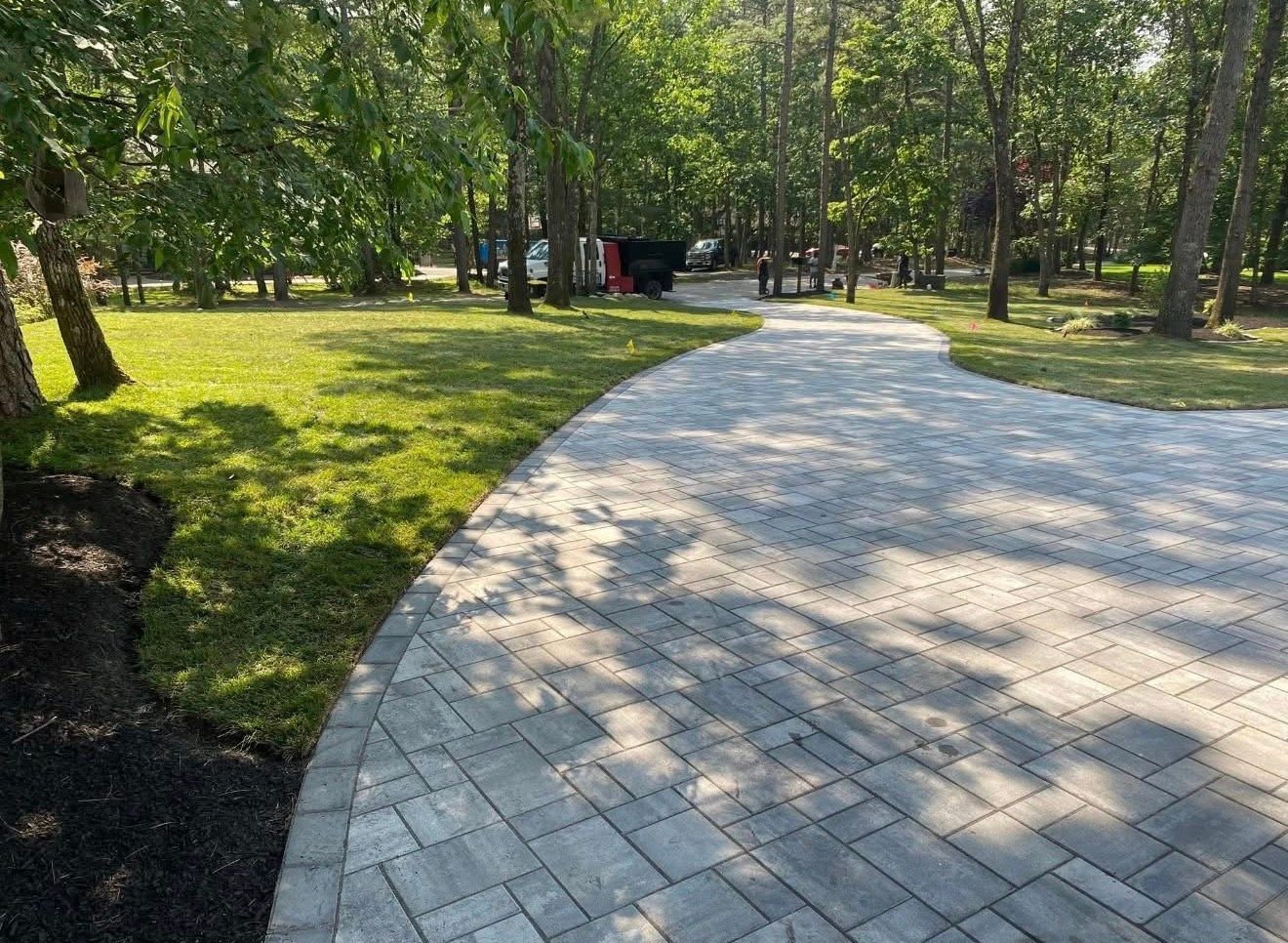 A light-colored stone paver driveway curves through a green, tree-filled yard on a sunny day.
