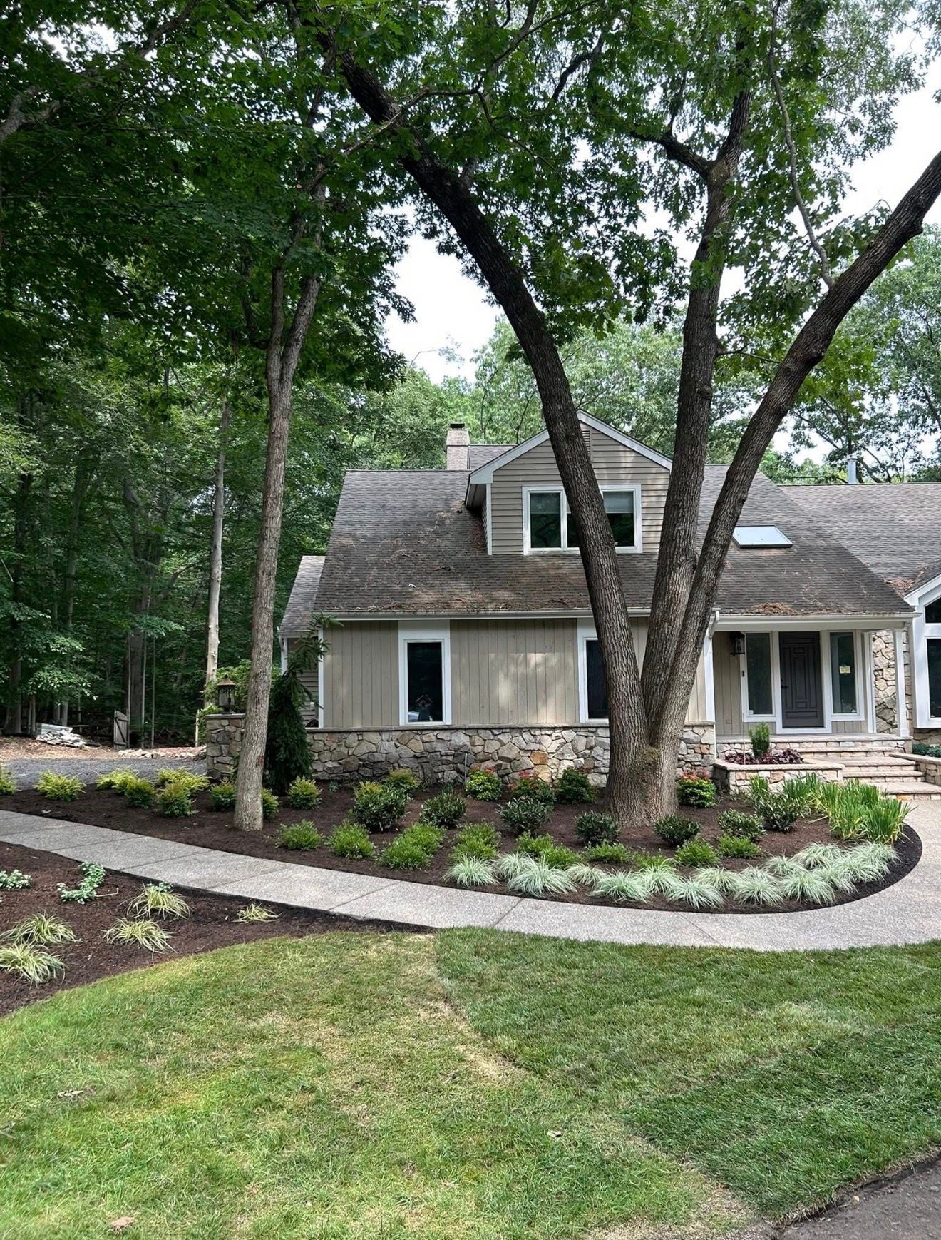 A tan-sided house with stone trim and a gray shingled roof, set behind a paved walkway and landscaped garden beds.