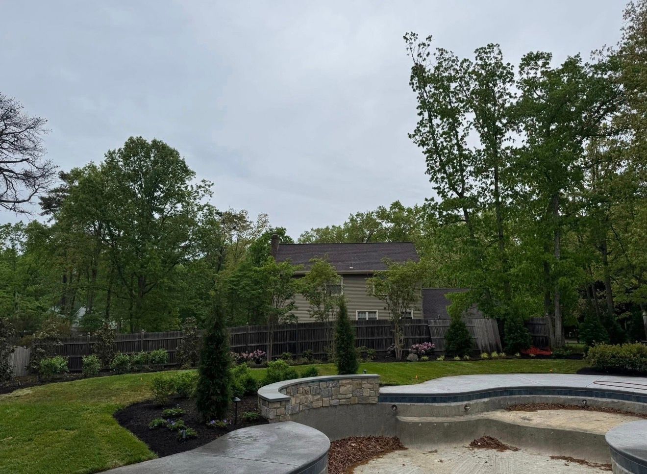 A backyard with a stone fire pit, landscaped beds with small evergreens, a wood fence, and trees under a cloudy sky.