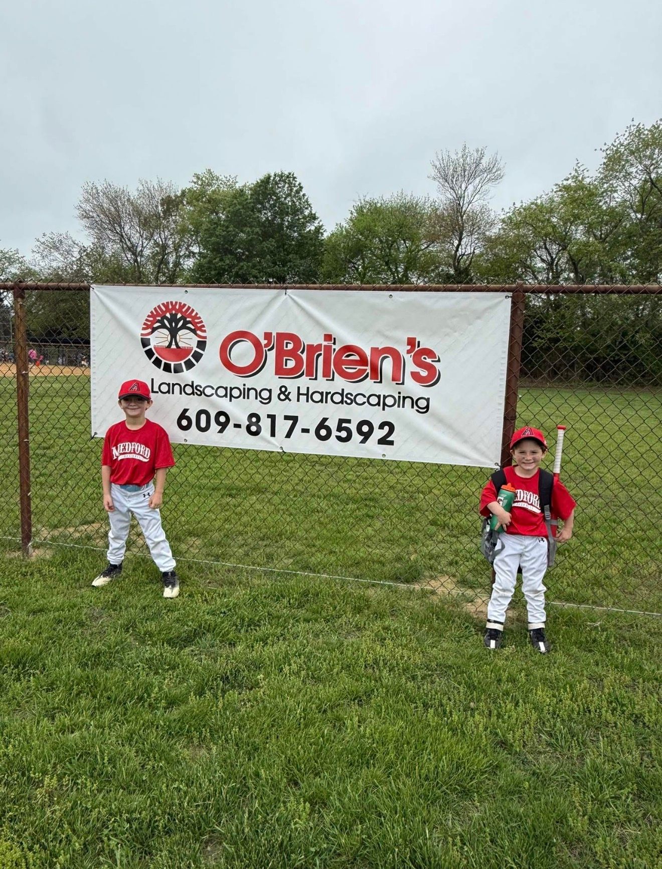 Two children in red and white baseball uniforms stand in front of a sponsor banner for O'Brien's Landscaping on a field.
