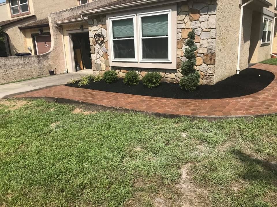 A brown brick walkway curves in front of a house with stone accents, dark mulch, and a manicured spiral shrub.