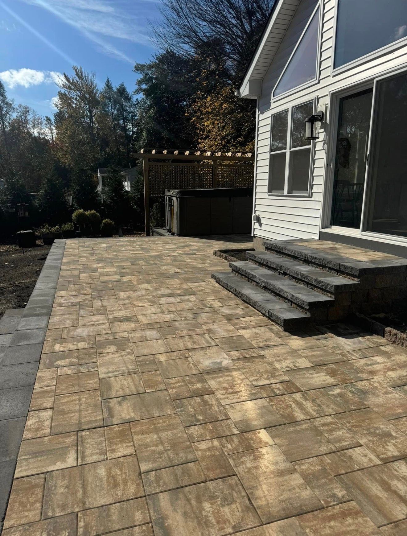 A paved patio with stone steps leading to a white house, surrounded by trees and a pergola in the backyard.