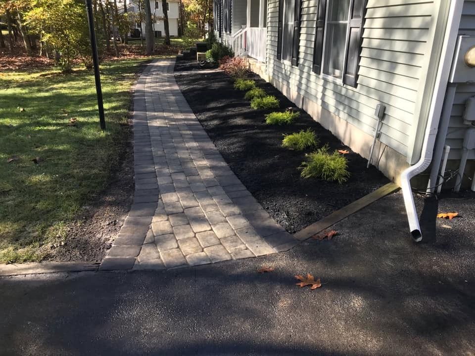 A paved stone walkway leads from an asphalt driveway toward a light-colored house with dark mulch beds and small shrubs.