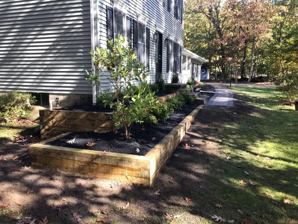 A terraced garden bed with a wooden retaining wall and fresh dark mulch in front of a house with grey siding.