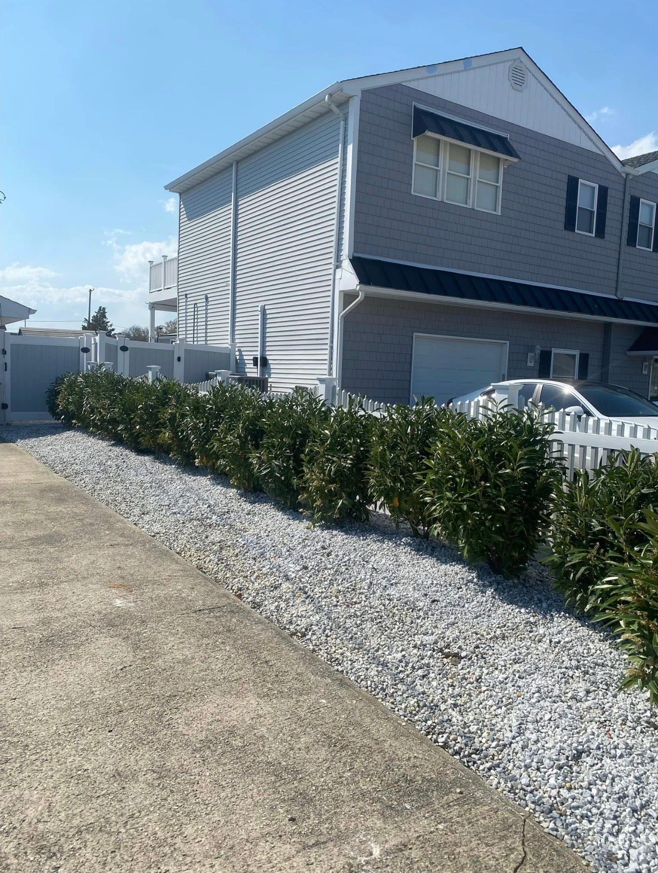 A gray two-story house with a white picket fence, gravel landscaping, and a row of green shrubs along a concrete driveway.