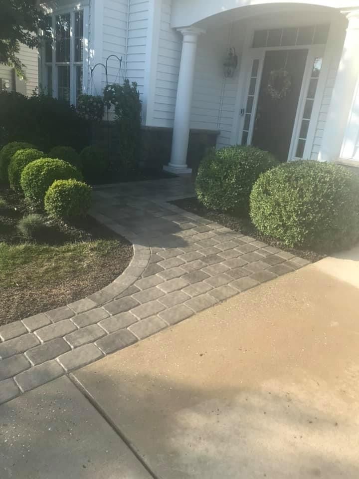 A paved stone walkway leads from a concrete driveway to the white front porch of a house with rounded green shrubs.