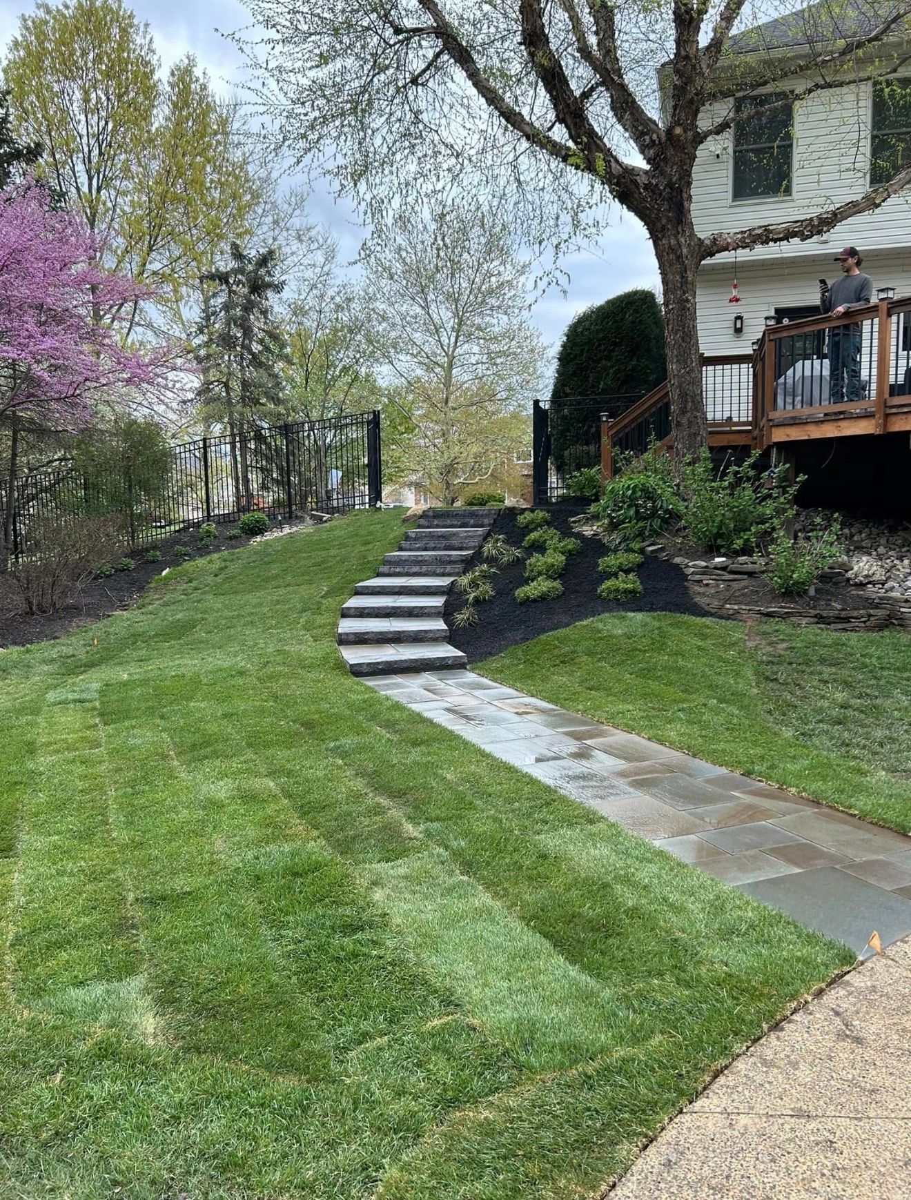 A flagstone walkway leads to stone steps ascending a grass-covered slope toward a house with a wooden deck.