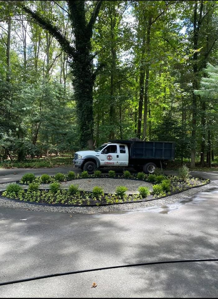 A white dump truck parked behind a small, circular garden bed with green shrubs in a paved, tree-lined driveway.