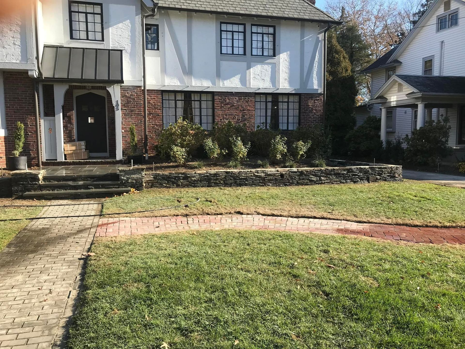 A Tudor-style house with brick and white trim, featuring a new stone retaining wall and a red brick curved walkway.