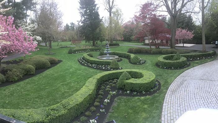 A formal garden features curved, manicured boxwood hedges, a small fountain, vibrant spring trees, and a cobblestone path.