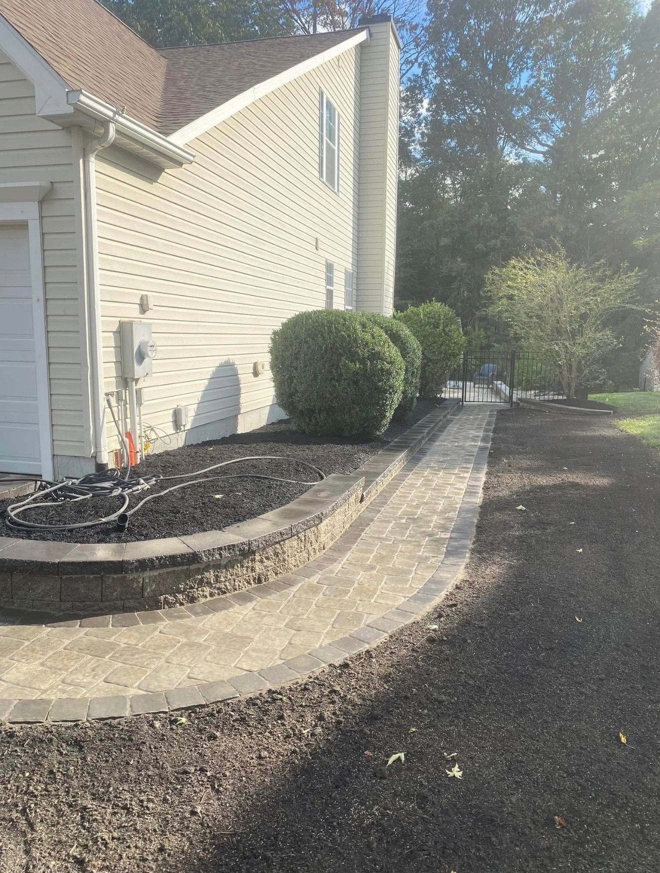 A curved paver walkway leads alongside a beige house with a stone retaining wall and rounded shrubs.
