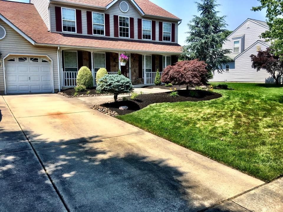 A tan suburban house with a paved driveway, green lawn, and manicured shrubs on a sunny day.