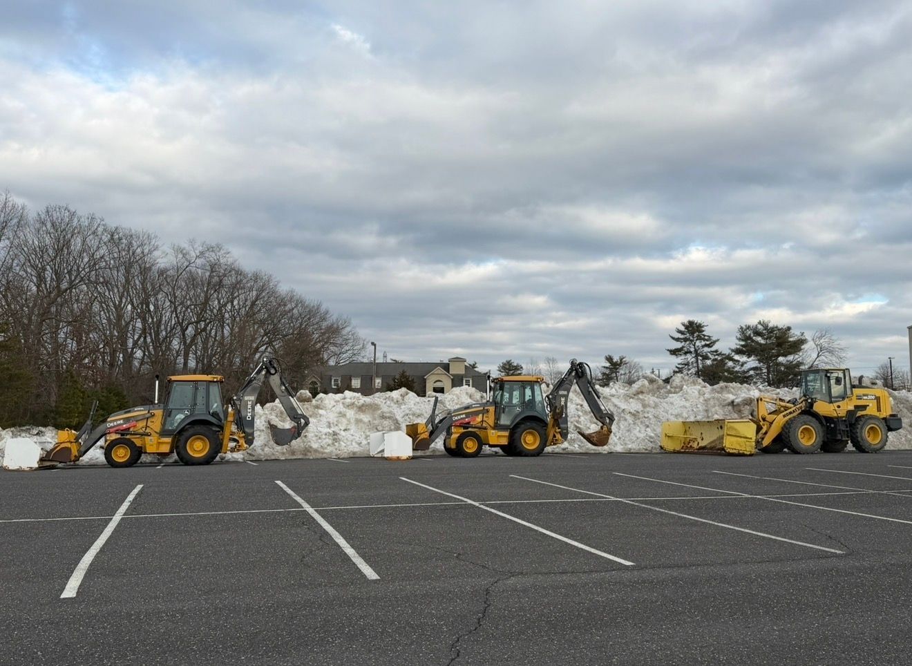 Three yellow construction vehicles parked in a row in a paved lot in front of a large pile of snow.
