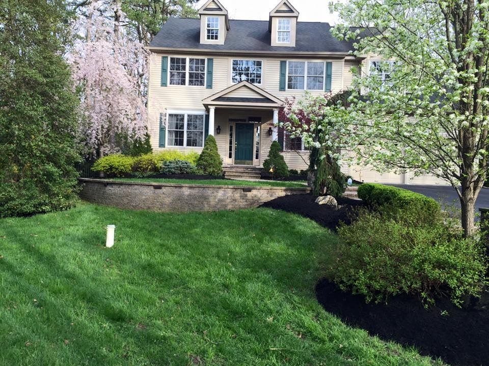 A two-story house with light beige siding and dark shutters behind a tiered stone retaining wall and landscaped yard.
