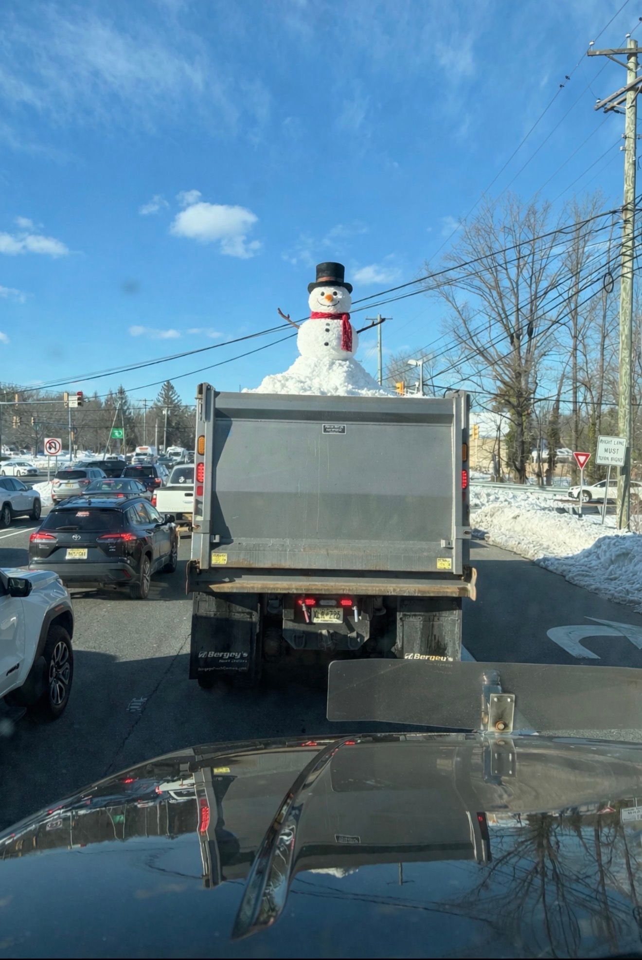 A snowman wearing a top hat and red scarf sits in the back of a dump truck idling in traffic on a sunny winter day.