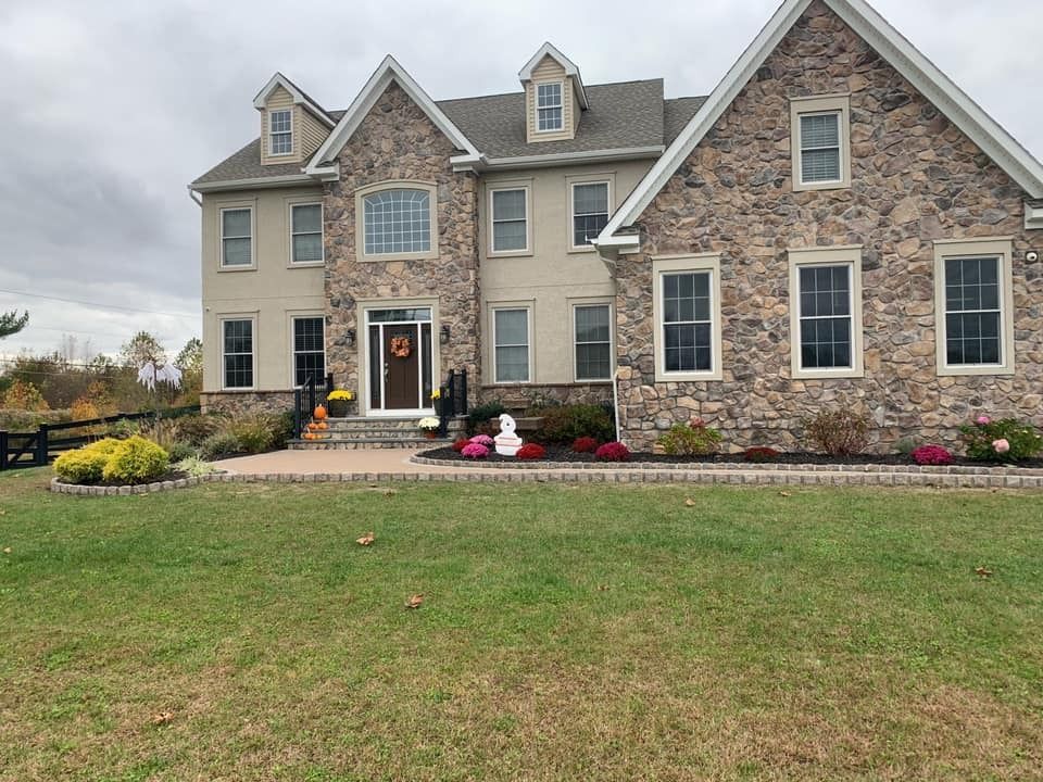 A two-story stone and beige house with a large front lawn under a cloudy autumn sky, featuring seasonal landscaping.