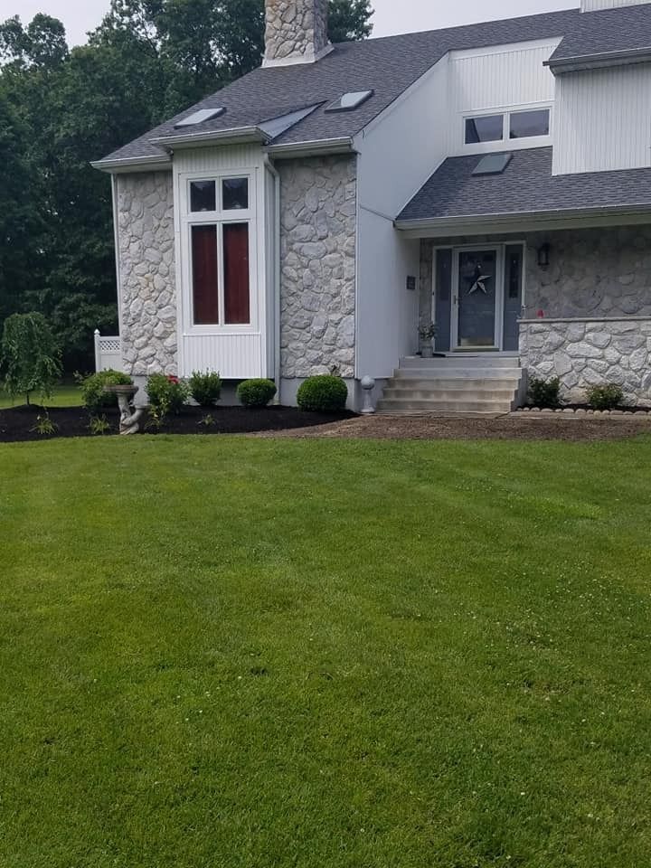 A two-story house with stone masonry and white siding, featuring a front lawn and a gray shingled roof under a cloudy sky.