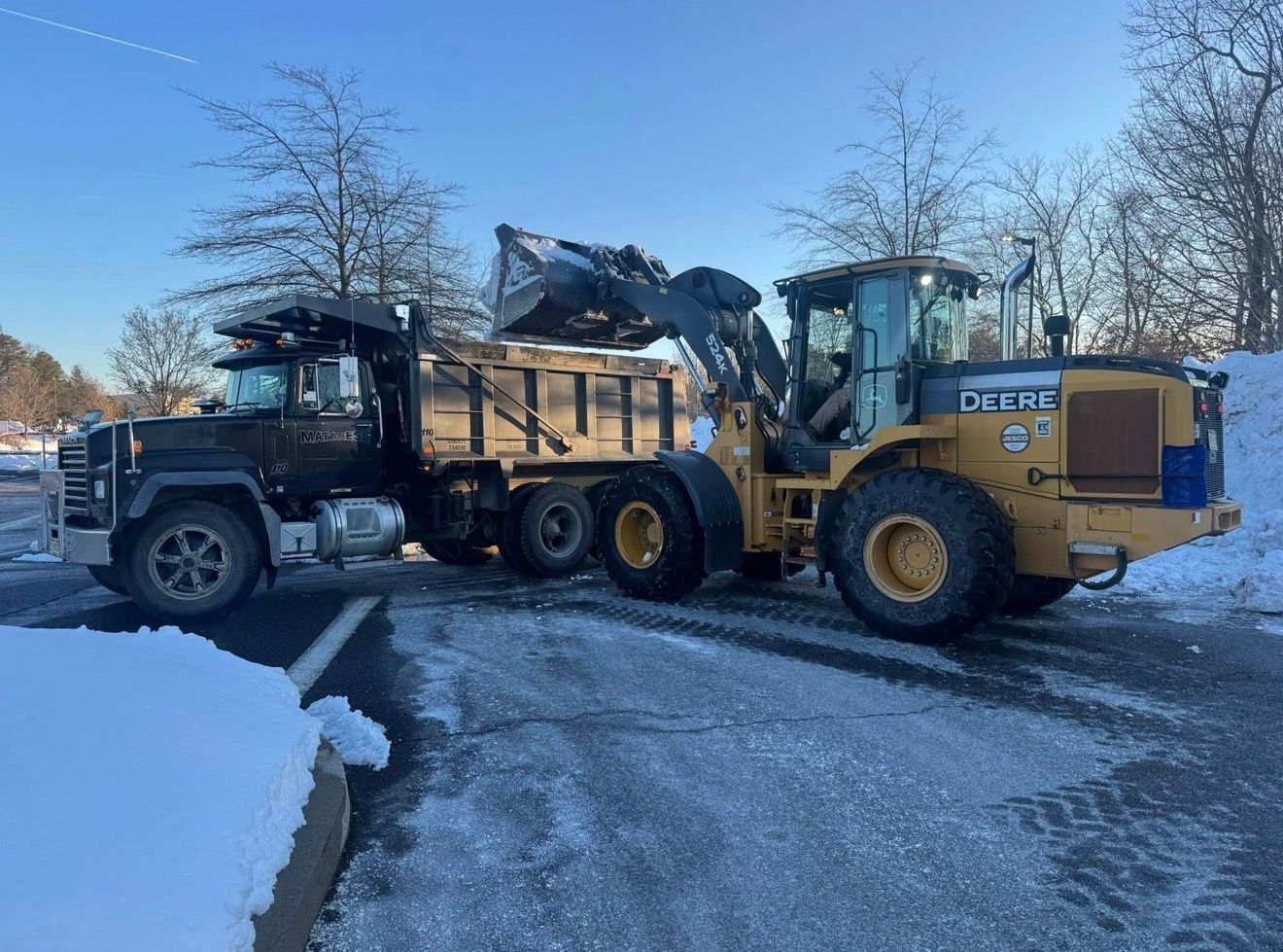 A yellow John Deere wheel loader dumping snow into the bed of a black dump truck in a snowy parking lot.