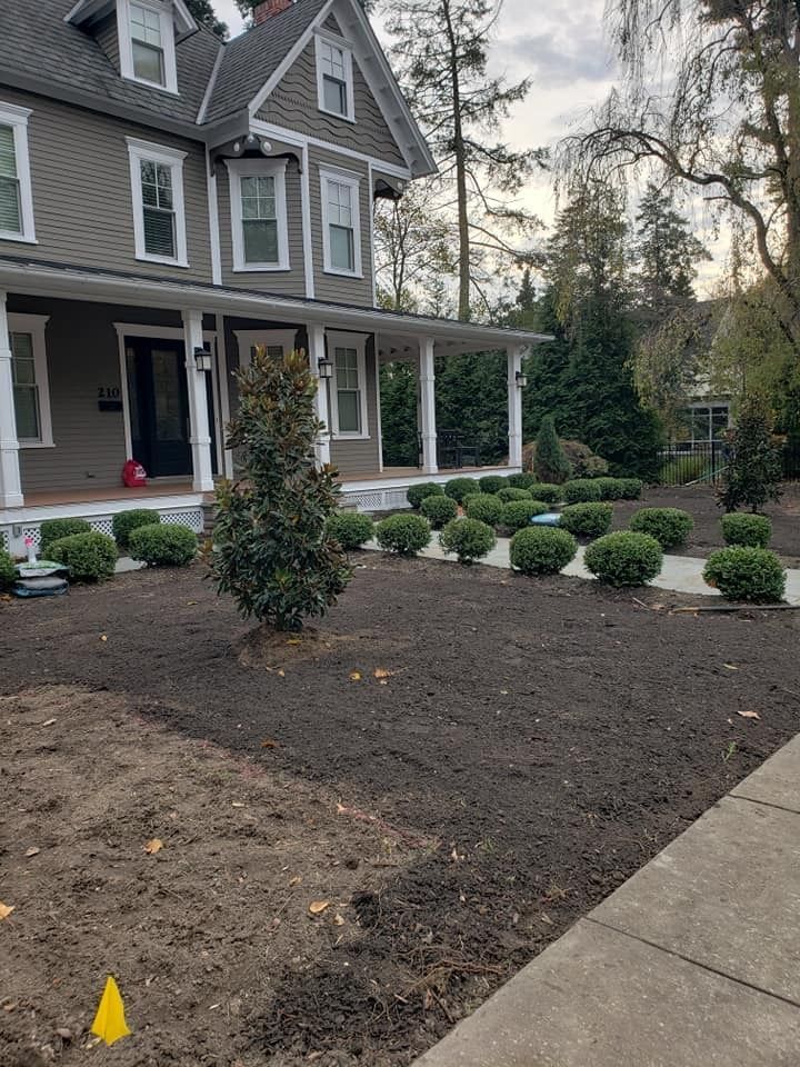 A gray, multi-story house with a white wrap-around porch, featuring fresh mulch and new shrubs planted in the front yard.