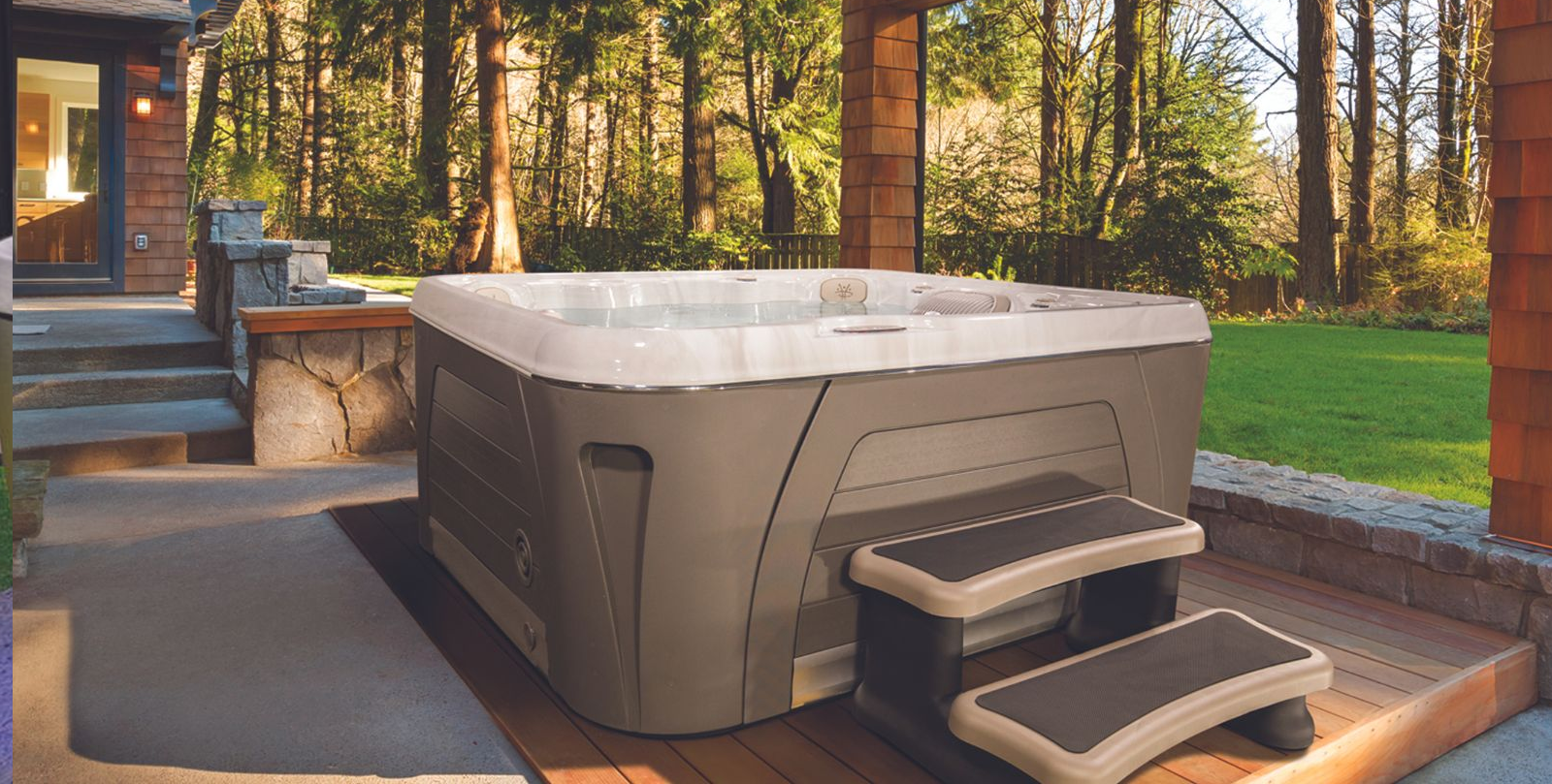 Outdoor gray hot tub on a patio with steps, set beside a brick wall and trees at sunset