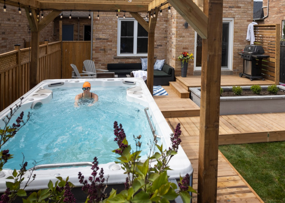 Person swimming in a small backyard pool beside a brick patio and wooden deck pergola.