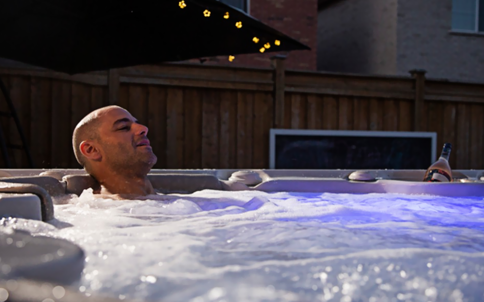 Person relaxing in a hot tub at dusk, with string lights glowing overhead.