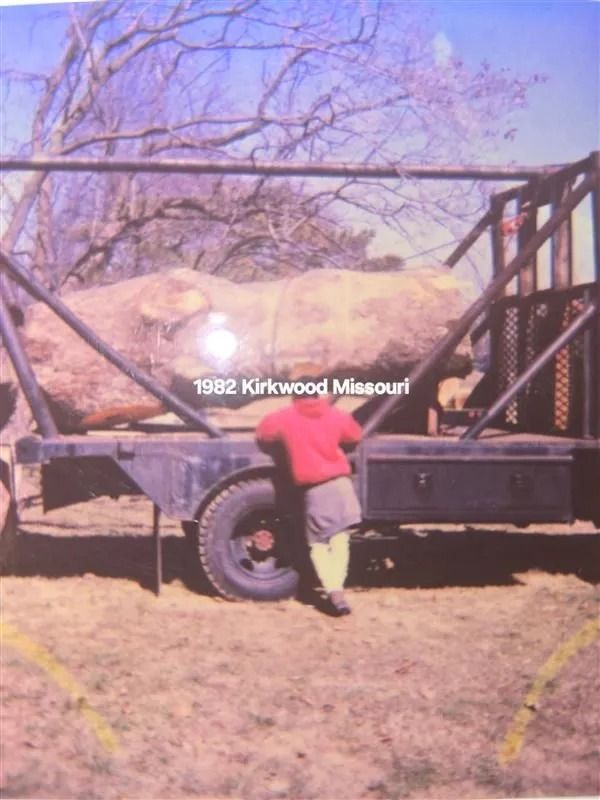 Boy in red shirt leans against a trailer holding a large log. 1982 Kirkwood, Missouri.