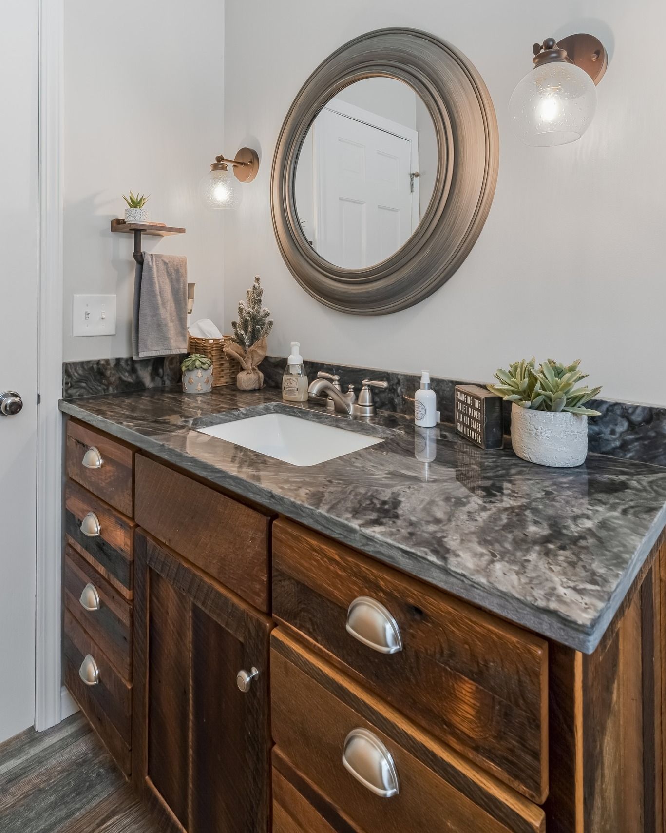 Bathroom vanity with dark wood cabinets, gray countertop, round mirror, and sconces.